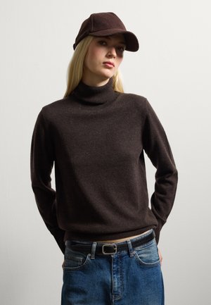 Maglione a collo alto marrone realizzato in materiale morbido, abbinato a un berretto scuro. Il modello indossa jeans blu e una cintura nera, posando di fronte a uno sfondo neutro.