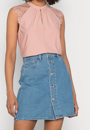 Femme portant un chemisier rose sans manches avec des épaules en dentelle et une fente à l'avant, associée à une jupe en denim bleu à boutons devant.
