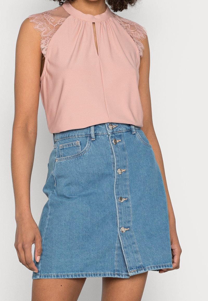 Femme portant un chemisier rose sans manches avec des épaules en dentelle et une fente à l'avant, associée à une jupe en denim bleu à boutons devant.