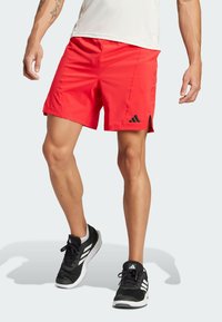 Shorts de sport rouges en tissu léger avec un logo Adidas noir et des fentes sur les côtés pour faciliter les mouvements, associés à des baskets noires.