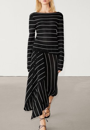Femme portant un haut noir à manches longues et une jupe asymétrique avec des rayures blanches, ainsi que des sandales noires à brides, debout sur un sol gris.