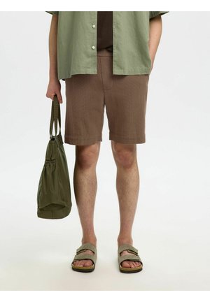 Muž nosiaci hnedé textúrované šortky, zelenú rozopnutú košeľu, držiaci olivovo zelenú tašku a béžové sandále na bielom pozadí.