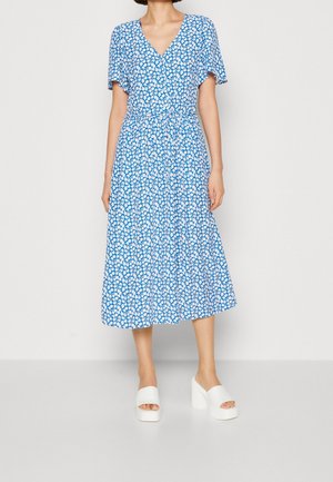 Frau trägt ein blaues Midikleid mit weißem Blumenmuster und kurzen Ärmeln, kombiniert mit weißen Plateausandalen mit hohen Absätzen.