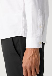 Camisa branca de manga longa com punho abotoado, apresentando uma textura suave e um rótulo lateral perto da bainha. Usada com calças pretas.