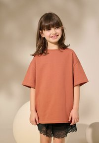 Rustrød oversized T-shirt med korte ærmer, parret med sorte shorts med blondekant. Glat tekstur, afslappet pasform.
