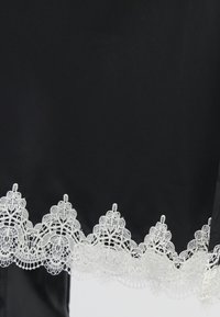 Tissu noir avec un bord en dentelle blanche présentant des motifs floraux complexes le long du bord inférieur. La texture lisse contraste avec la dentelle.