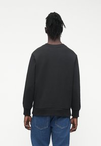 Muž s krátkými dredy, oblečený v černé mikině s dlouhými rukávy a modrých džínách, stojící zády k nám na jednobarevném pozadí.