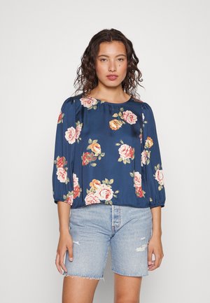 Blusa floral em tecido azul-marinho com mangas bufantes de três quartos. Apresenta um decote redondo e grandes padrões de rosas cor-de-rosa e laranja. Combinada com calções de ganga.