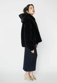 Veste oversized en fausse fourrure noire avec capuche, associée à une longue jupe noire et des sandales dorées à lanières. Texture lisse et coupe décontractée.