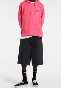 Sudadera rosa con logo azul de Vans, pantalones cortos de mezclilla negros y sneakers negros de Vans. El modelo lleva calcetas cortas negras con logo blanco.