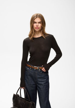 Jeune femme aux cheveux lisses châtain clair, portant un haut noir à manches longues, un jean bleu, une ceinture marron, tenant un grand sac à main noir.