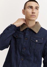 Denim jas in donkerblauw met een tan bontgekraagde kraag, knopen als sluiting en twee borstzakken met knopen. Heeft een ontspannen pasvorm.