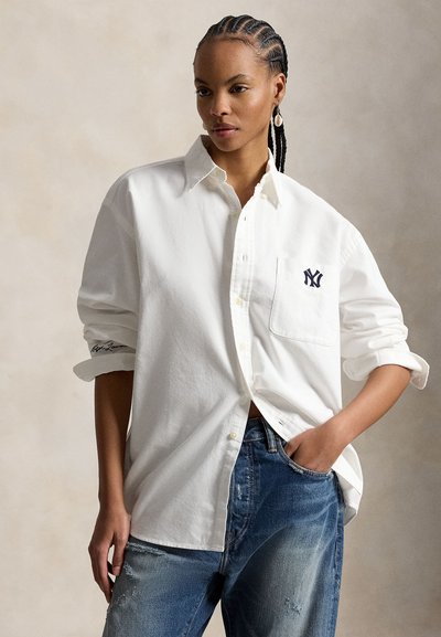 Frau mit geflochtenen Haaren, die ein übergroßes weißes Hemd mit aufgekrempelten Ärmeln und blaue Jeans trägt, mit einer Hand in der Hosentasche stehend.