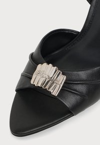 Sandale en cuir noir à bout ouvert avec une boucle décorative argentée présentant un détail de signature gravé sur la bride avant.