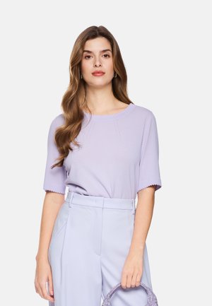 Frau mit langen welligen Haaren, die ein helllila kurzärmeliges Oberteil und passende hoch taillierte Hose trägt, hält eine lila strukturierte Handtasche.