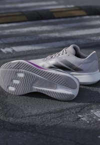 Hellgraue Sportschuhe mit lila Akzenten, ein Schuh mit der Sohle nach oben, der Profil-Details zeigt, auf strukturiertem Asphalt nahe einem Zebrastreifen platziert.