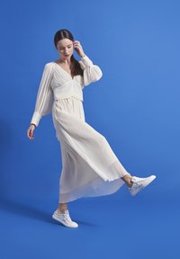 White pleated dress with long sleeves and a V-neck, paired with white sneakers, against a solid blue background. Light, flowing texture.