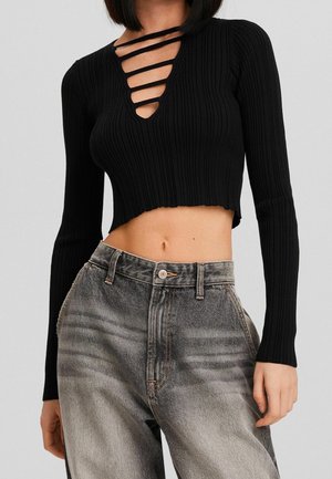 Femme portant un crop top noir côtelé à manches longues avec des détails de lanières horizontales au niveau de la poitrine et un jean gris délavé taille haute.