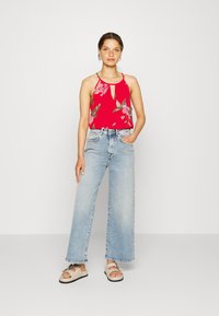 Top halter vermelho com estampado floral e decote em gota, combinado com jeans de perna larga azul-claro e sandálias beijas com detalhes decorativos.