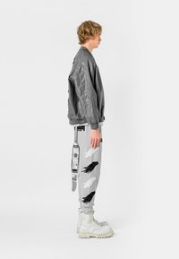 Chaqueta bomber gris con un corte relajado, combinada con pantalones gris claro que presentan patrones de plumas en blanco y negro y acentos gráficos detallados. Botas de combate blancas.