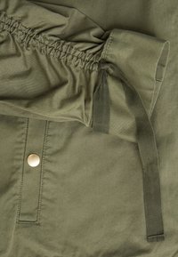 Olivgrön tyg med samlad detalj, matchande knytning och en rund metallknapp. Slät textur och synlig söm på tyget.