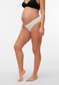 Έγκυος γυναίκα που φοράει μαύρο σουτιέν και λευκά εσώρουχα εγκυμοσύνης, στέκεται ξυπόλυτη στο ένα πόδι μπροστά σε απλό λευκό φόντο.