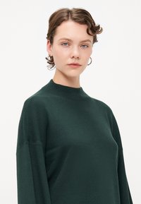 Mørkegrønn, strikket genser med høy hals og senkede skuldre. Stoffet ser mykt ut, med en jevn tekstur og minimale detaljer.