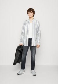 Een geknoopte, button-up overhemd met blauwe en witte verticale strepen. Draagt over een witte tanktop, gecombineerd met donkere jeans en lichte sneakers.