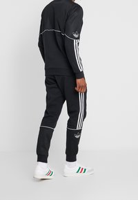 Persona vistiendo un chándal negro de Adidas con rayas blancas y logotipos de trébol, combinado con zapatillas blancas que tienen rayas verdes y rojas.