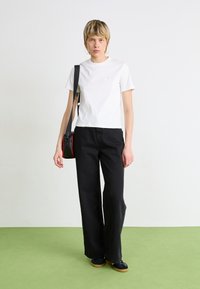 Hvid kortærmet t-shirt med afslappet fit; sammen med højtaljede sorte brede bukser. Sort crossbody taske med røde detaljer. Grøn gulvbaggrund.