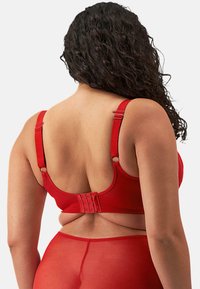 Sujetador rojo con tirantes ajustables, textura suave, ganchos en la parte posterior y una braguita transparente a juego. Enfoque en el diseño funcional y la comodidad del ajuste.