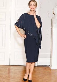 Robe marine avec un voile fluide et des garnitures en sequins, présentant une silhouette droite et une longueur au genou. Des escarpins noirs complètent le look.