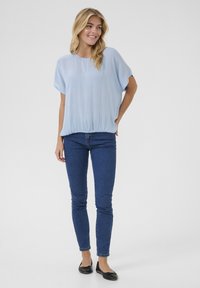 Blouse bleu clair à manches courtes avec une coupe décontractée, ourlet élastique, portée avec un jean skinny en denim foncé et des ballerines noires.
