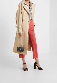 Beige trenchcoat, geruite blouse, koraalkleurige tailor made broek, zwarte sandalen met bandjes, geruite mini-tas met ketting. Heeft een klassiek ontwerp en levendige kleuren.
