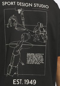 Zwart t-shirt met witte grafische illustraties van basketbalspelers en tekst. Toont dynamische poses en acties in een sportthema.
