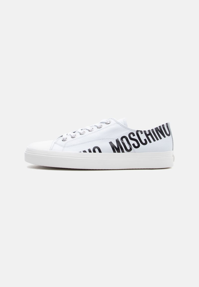 Baskets basses blanches avec le logo "MOSCHINO" en noir sur le côté et des lacets blancs, posées sur un fond gris clair uni.