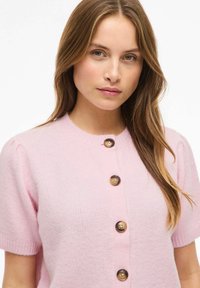 Svijetloružičasti pleteni kardigan s kratkim napuhanih rukavima i pet velikih smeđih dugmadi. Mekana tekstura, okrugli izrez oko vrata i suptilan uzorak pletenja.