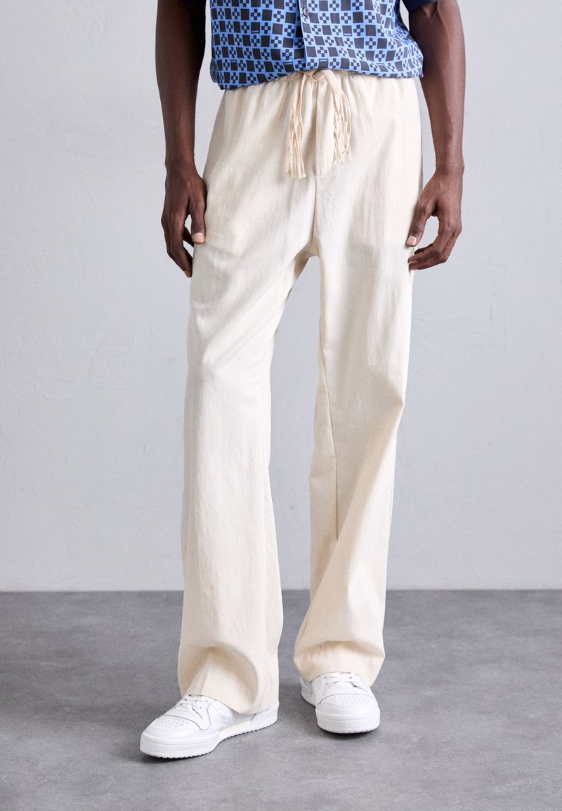 Pantalons amples de couleur crème en tissu doux, dotés d'une taille élastique et d'un cordon de serrage, associés à des baskets blanches.