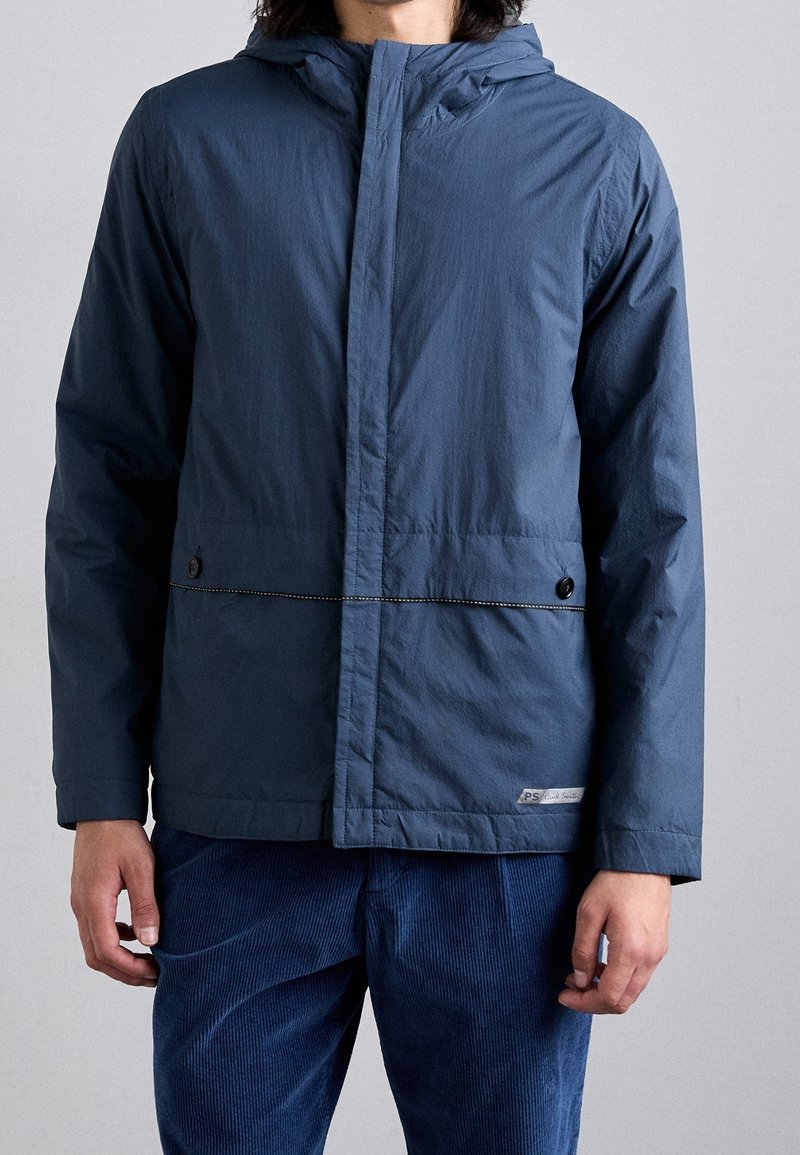 Homme portant une veste à capuche bleue avec des poches boutonnées et un pantalon en velours côtelé bleu assorti sur un fond uni.