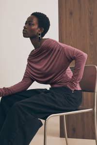 Långärmad, draperad topp i mauve tyg med rynkor, parat med lösa svarta byxor. Modellen bär svarta geometriska örhängen och sitter på en metallstol.