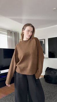 Orehasto rjav pulover iz pletenine z rebrastim izrezom in manšetami, kombiniran z ohlapnimi črnimi hlačami. Gladek teksturo, sproščen kroj, minimalističen dizajn.