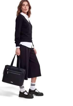 Donna con maglione nero, camicia bianca, pantaloni culotte neri, scarpe nere con calzini bianchi, che tiene una borsa a mano nera in pelle.