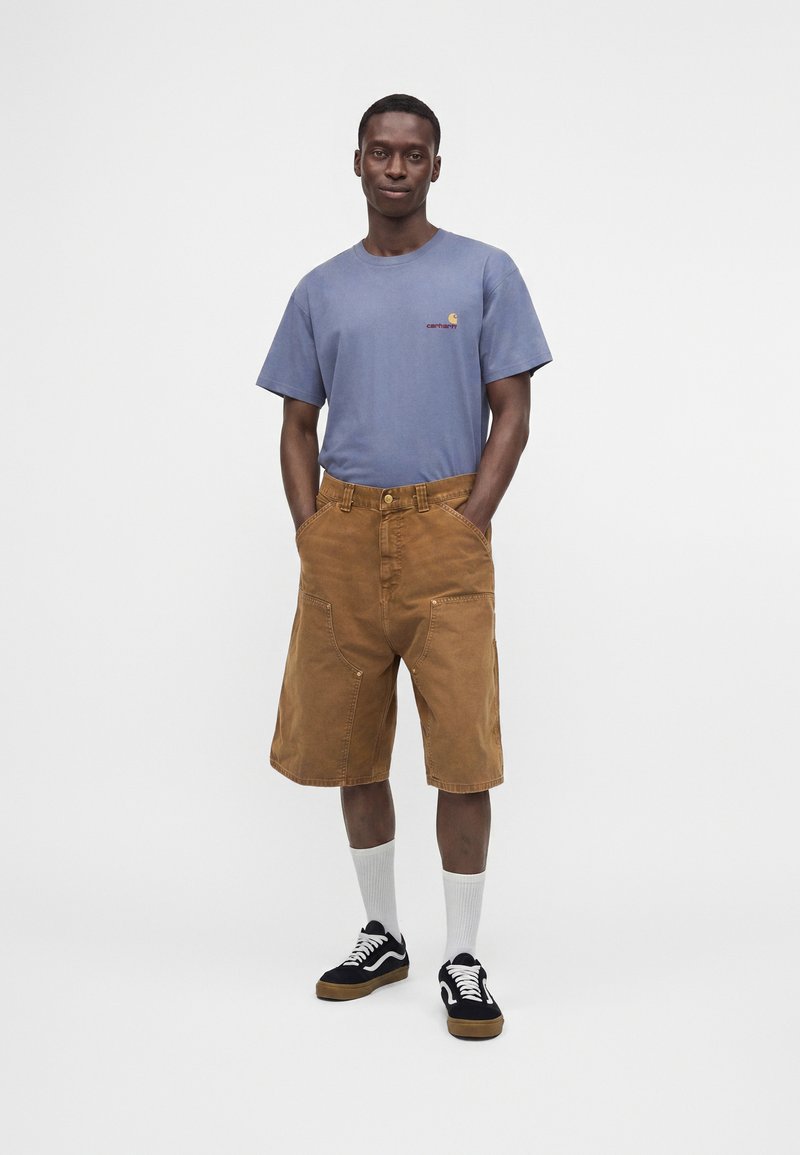 Joven de pie con las manos en los bolsillos, lleva una camiseta azul, pantalones cortos marrones hasta la rodilla, calcetines blancos y zapatillas negras sobre un fondo liso.