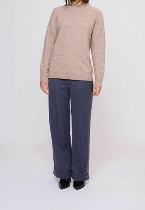 Persoon draagt een beige gebreide trui, marineblauwe wijde pijpen met krijtstreep en omgeslagen onderkant, en zwarte puntige schoenen tegen een effen achtergrond.
