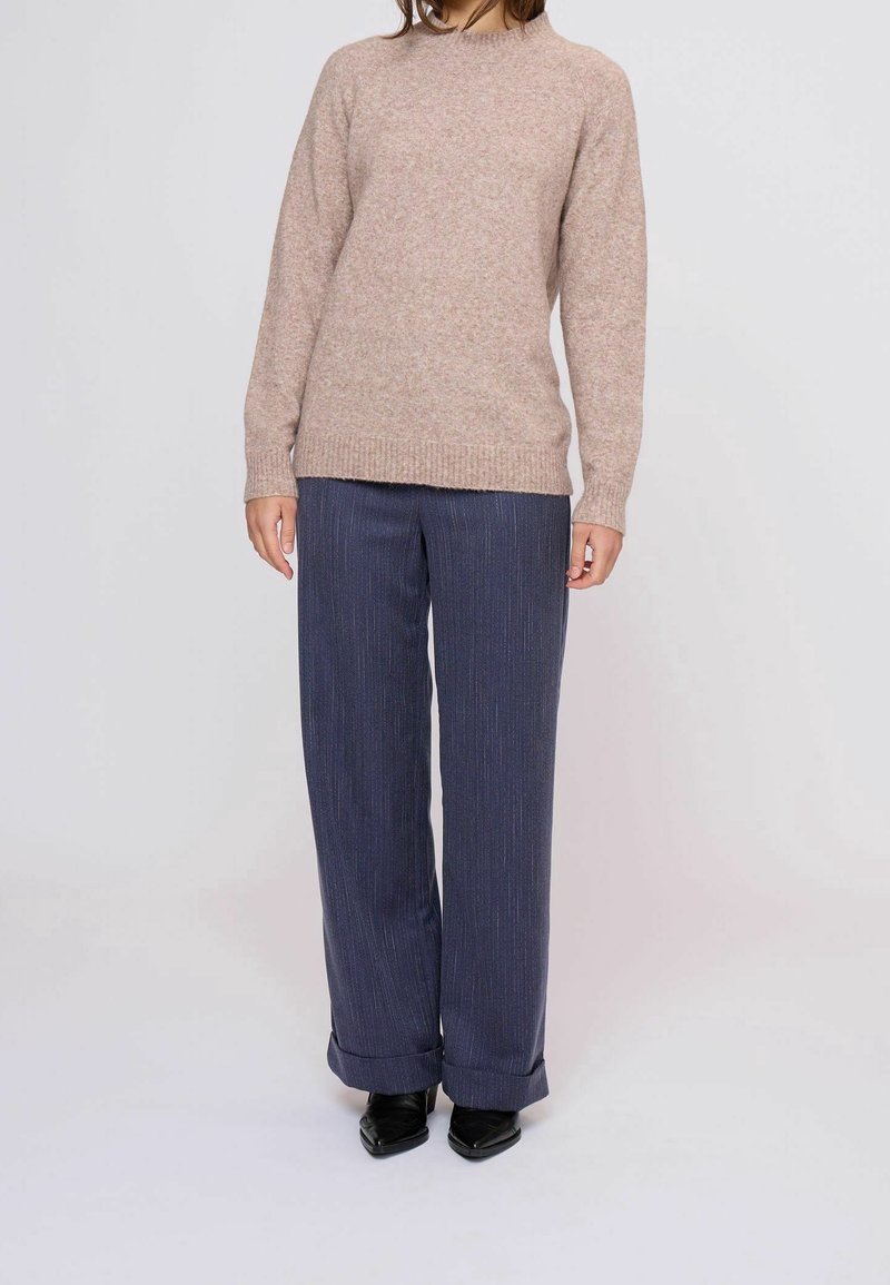 Personne portant un pull en maille beige, un pantalon large à rayures bleu marine avec revers au bas, et des chaussures noires pointues sur un fond uni.