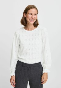 Camicia bianca a maniche lunghe in tessuto a perforazioni, scollo rotondo e polsini arricciati, abbinata a pantaloni grigi scuri sartoriali.