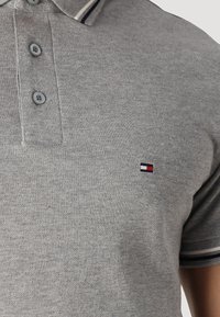 Šedé polo tričko vyrobené z texturované tkaniny, s knoflíkovou légou na tři knoflíky a malým vyšitým logem v modré, bílé a červené barvě.