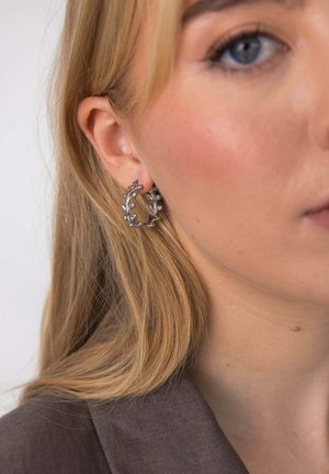 Nahaufnahme einer Frau mit blondem Haar, die silberne Creolen in Form von Zweigen mit kleinen Blättern trägt.