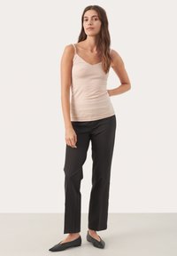 Lichtbeige tanktop met dunne banden, aansluitend design, gecombineerd met zwarte taillepantalon en donkere platte schoenen, weergegeven tegen een effen achtergrond.