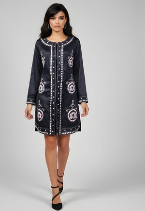 Mujer vistiendo un vestido negro hasta la rodilla con patrones florales bordados en blanco y tacones negros con tiras, de pie frente a un fondo gris liso.